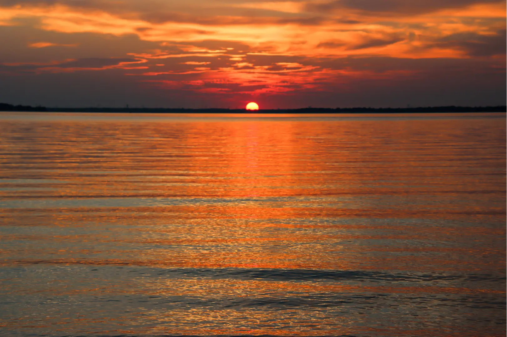 A vibrant sunset over Lake Lavon in Texas, with the sun partially set on the horizon and its warm orange and red hues reflecting across the calm water surface under a partly cloudy sky.