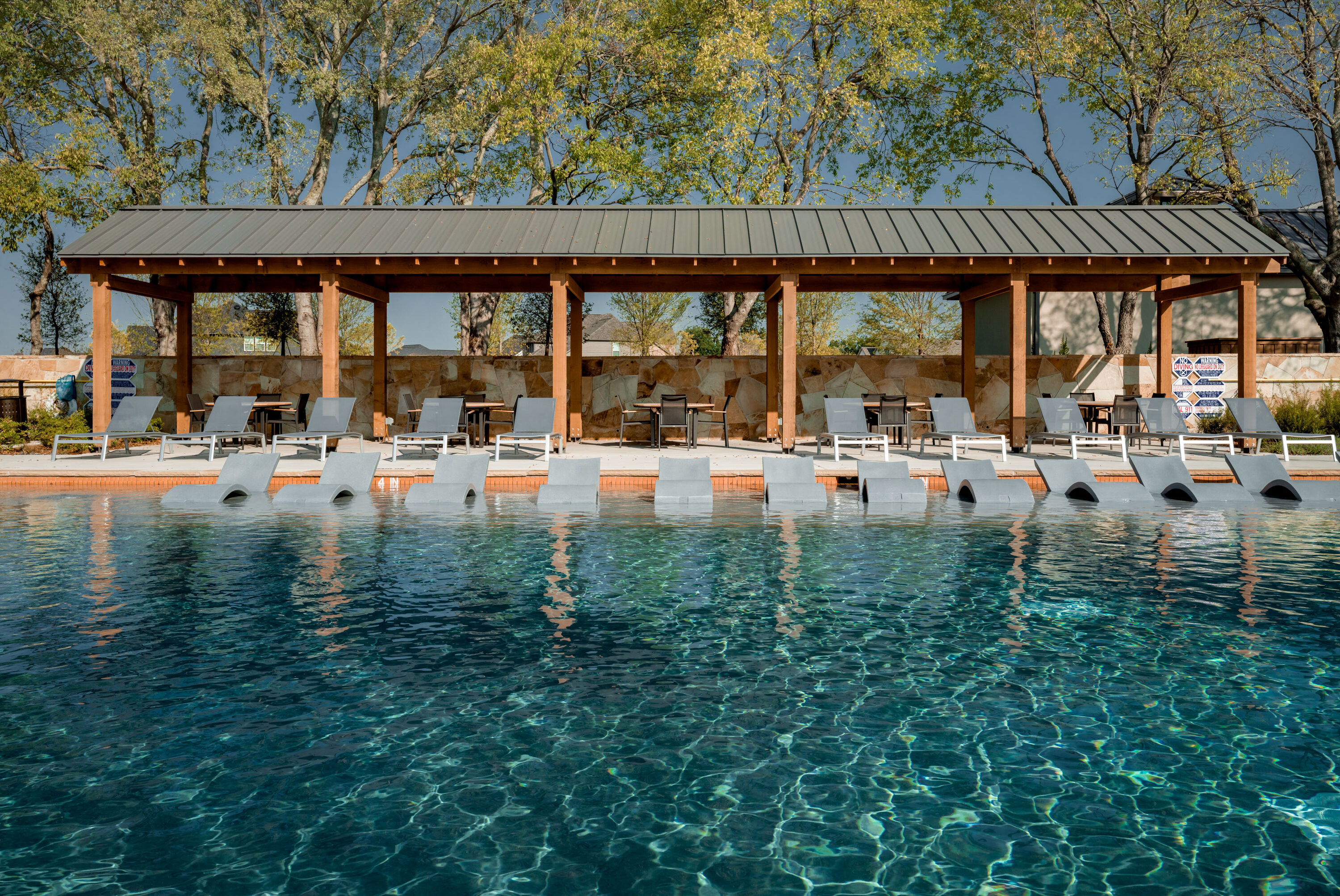 Resort-style swimming pool with in-water lounge chairs and shaded cabana seating surrounded by trees at Hillstead.