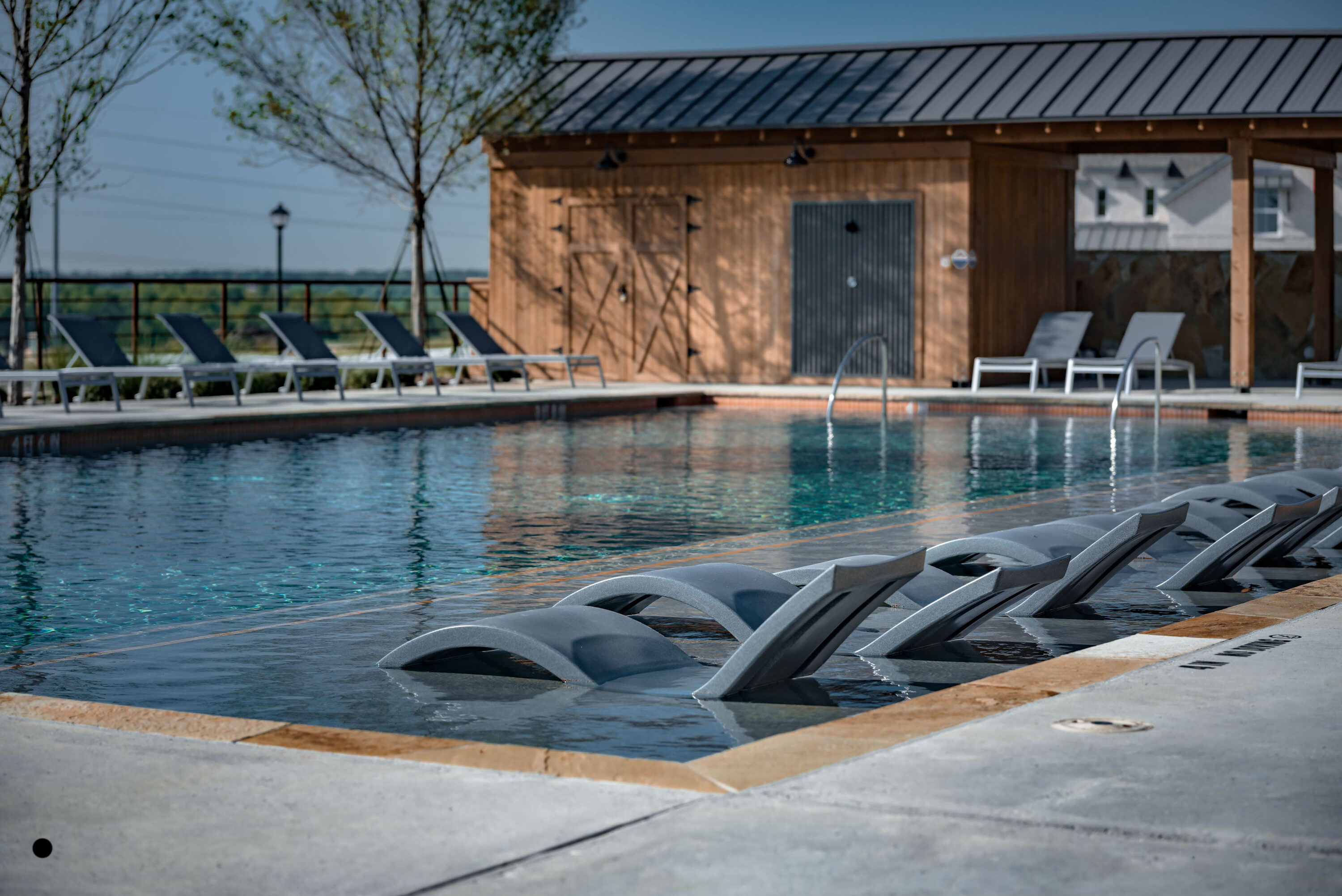 Outdoor swimming pool with in-water lounge chairs and sun deck seating beside a modern cabana at Hillstead.