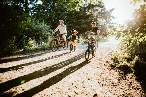 A family riding bicycles together on a sunlit dirt trail surrounded by trees, with two adults and two children enjoying a leisurely ride outdoors.