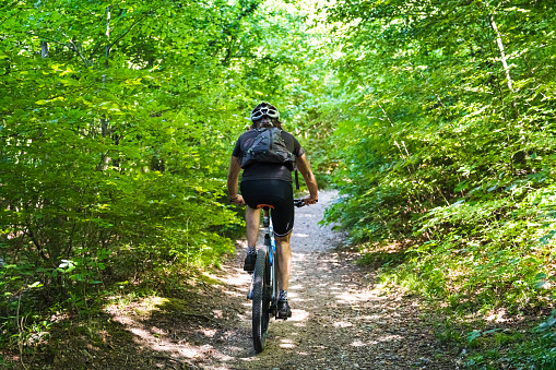 A cyclist wearing a helmet and backpack rides a mountain bike along a narrow dirt trail through a lush green forest on a sunny day.