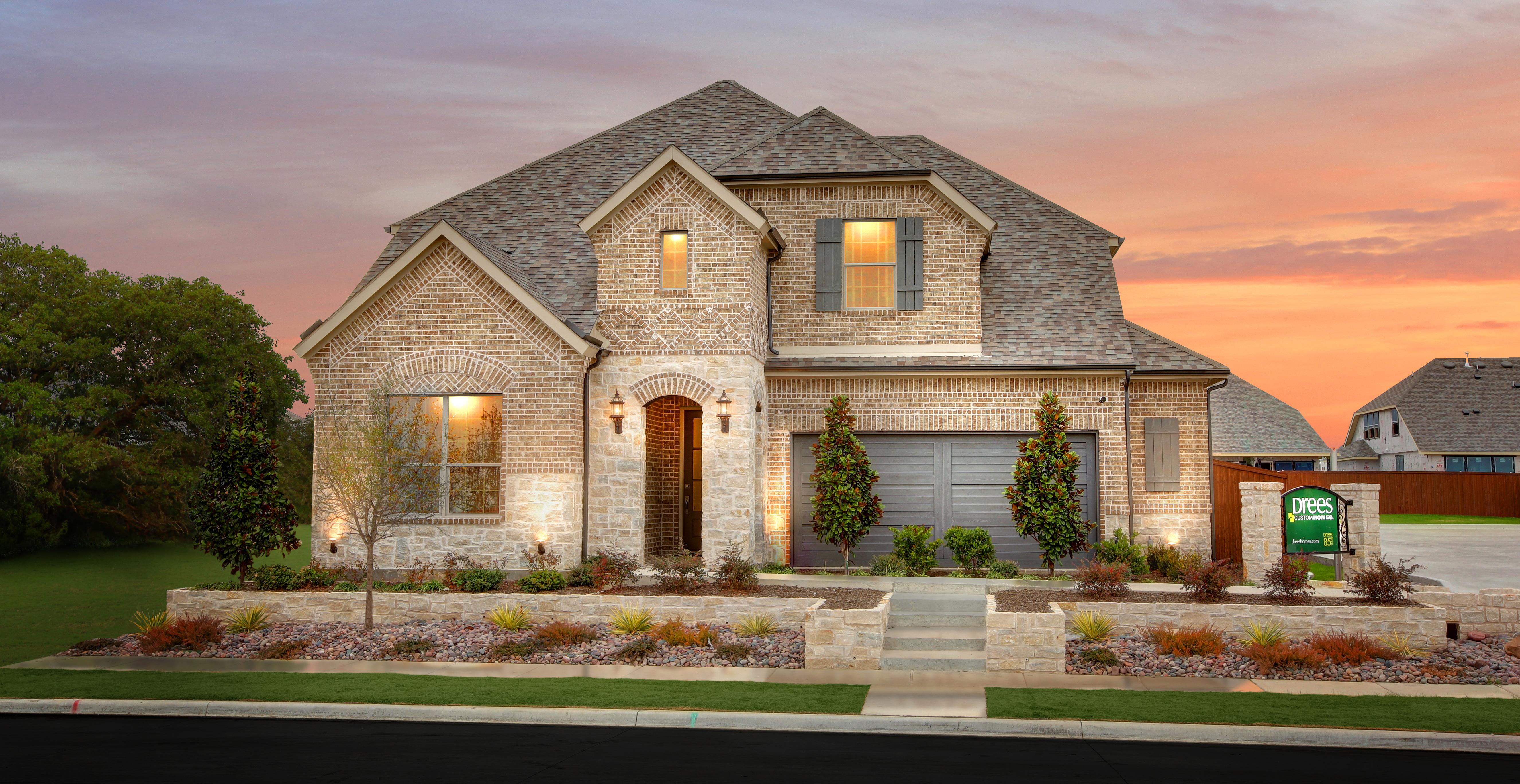 Front exterior of a two-story brick and stone home with landscaped yard, front steps, and attached garage, photographed at sunset.