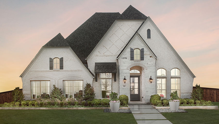 White brick new construction home with steep gabled roof, arched windows, manicured landscaping, and front walkway at sunset in a Texas residential community