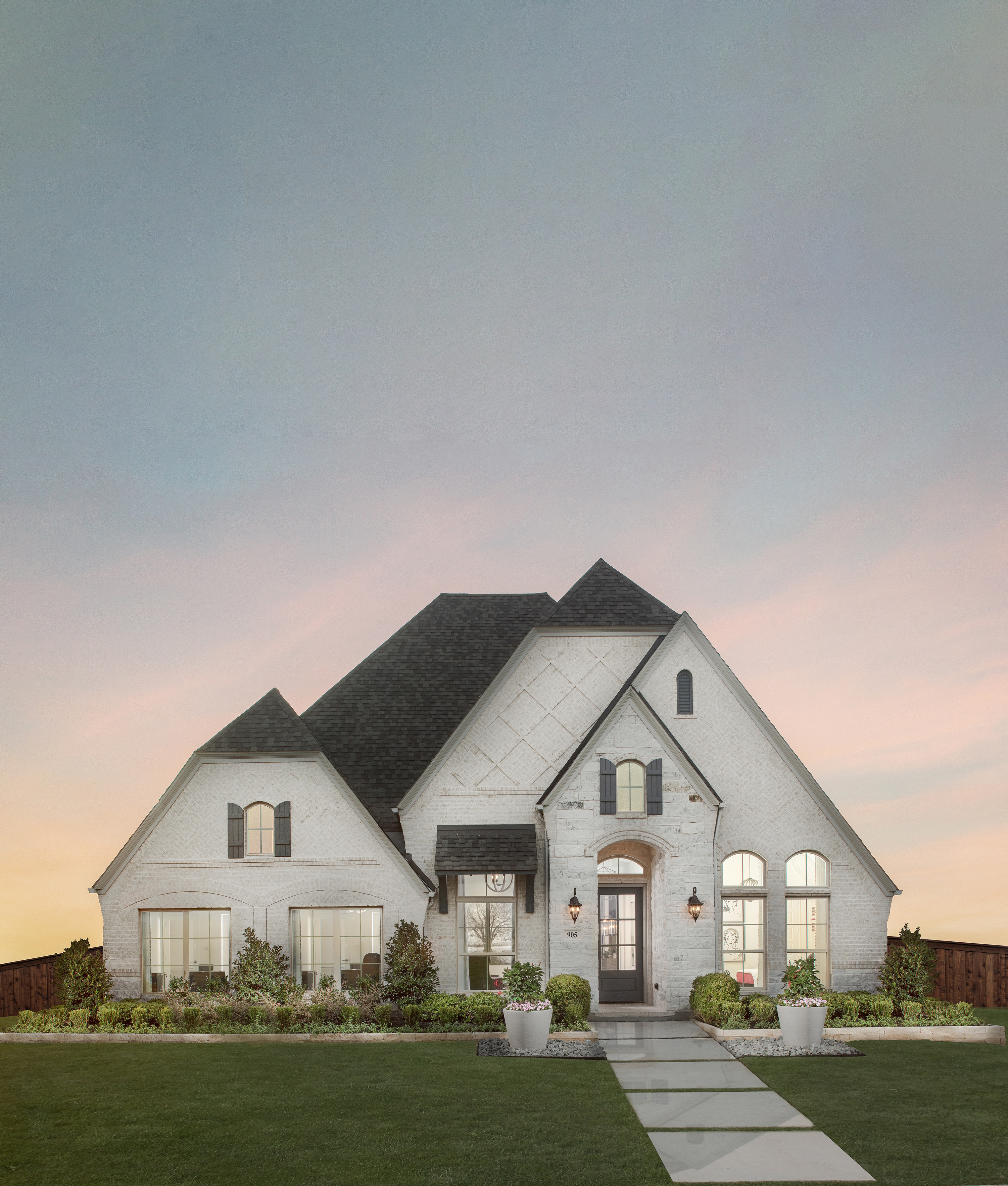 White brick new construction home with steep gabled roof, arched windows, manicured landscaping, and front walkway at sunset in a Texas residential community