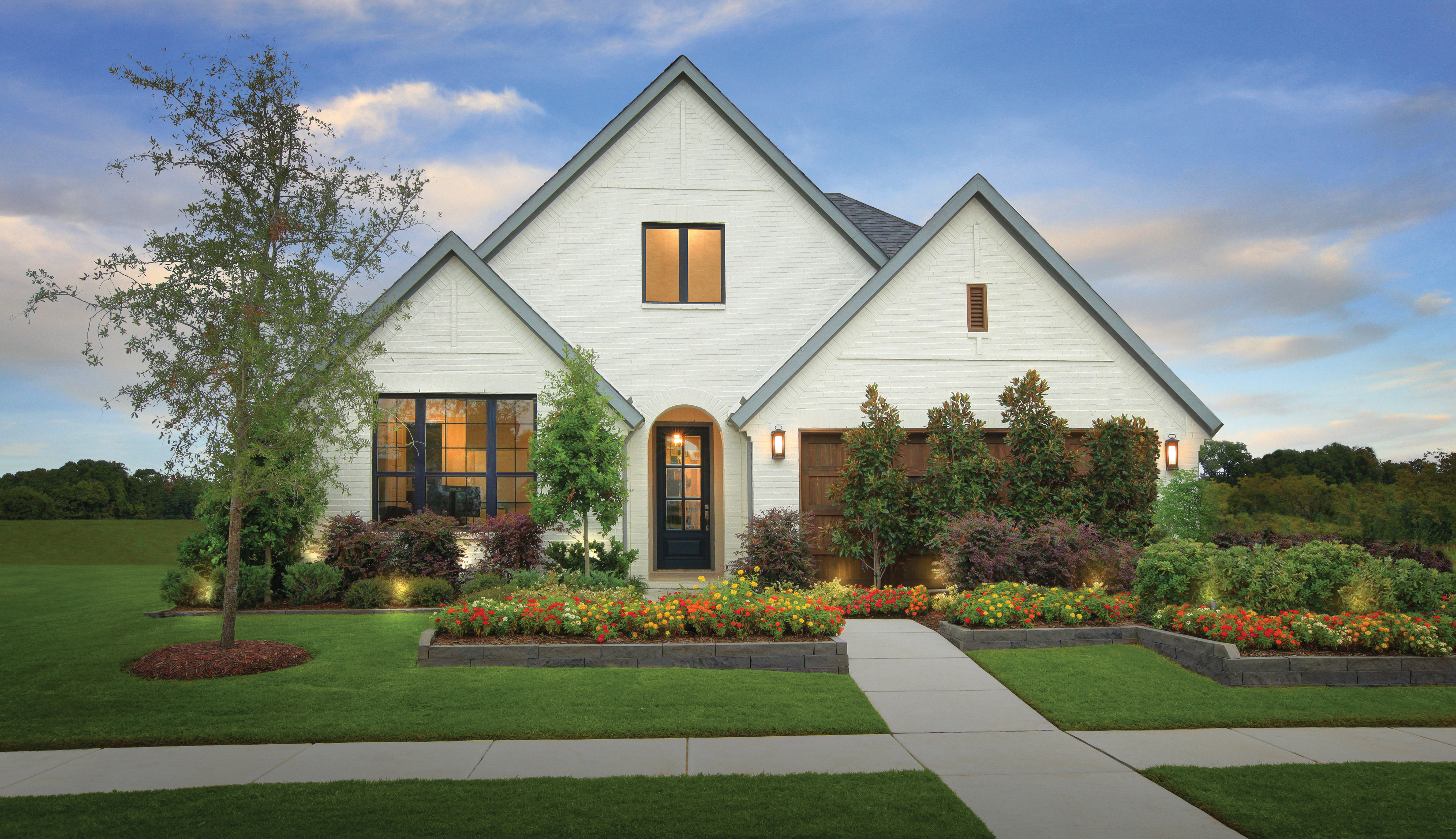 Modern white farmhouse with black windows, wood garage door, landscaped yard, and evening exterior lighting.